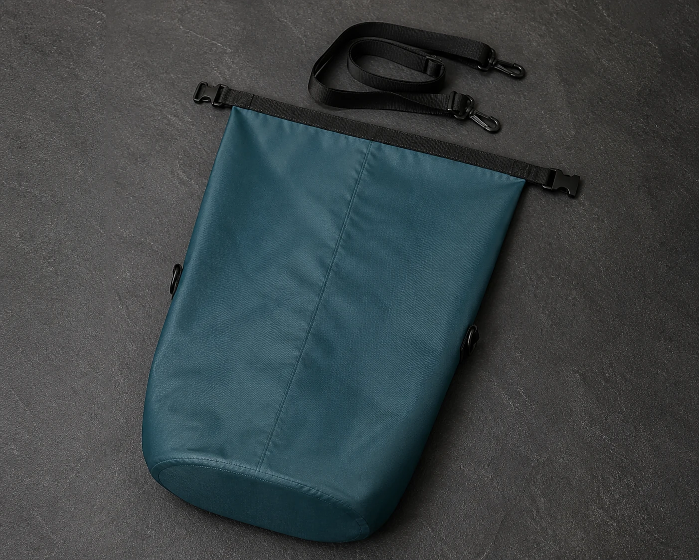 Single dark teal roll-top bag laid flat with its mouth open, buckle halves separated and shoulder strap detached above it
