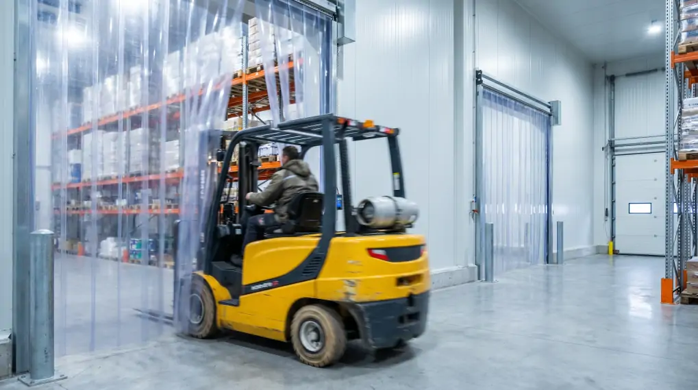 Industrial forklift driving through magnetic plastic vinyl curtains in cold storage warehouse showing hands-free operation