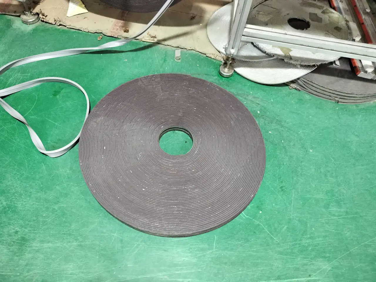 A coiled gray flexible edge material resting on a green workshop floor near additional long profiles and processing equipment.