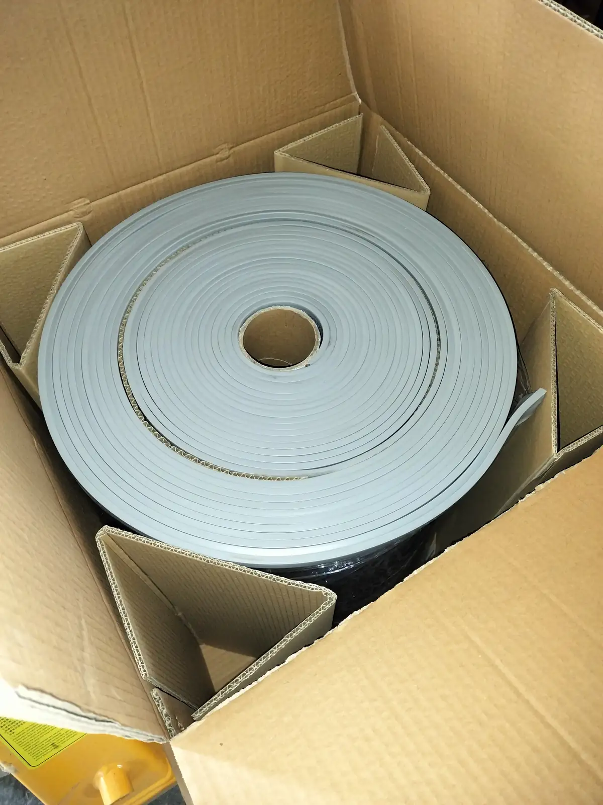A large coiled gray flexible strip packed inside a corrugated carton with folded cardboard separators.