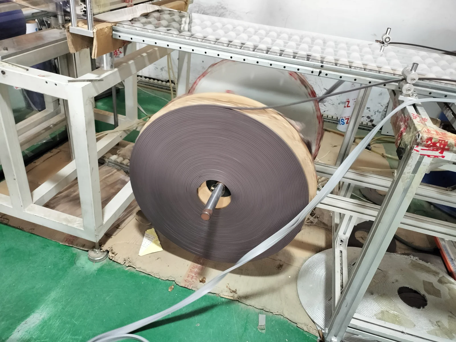 Large dark gray coiled profile mounted on a feed spindle with a narrow gray strip routed through workshop equipment.