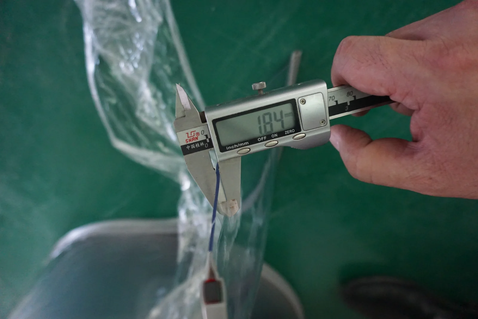 Handheld digital caliper applied to the edge of a clear flexible sample, with the jaws and display visible.